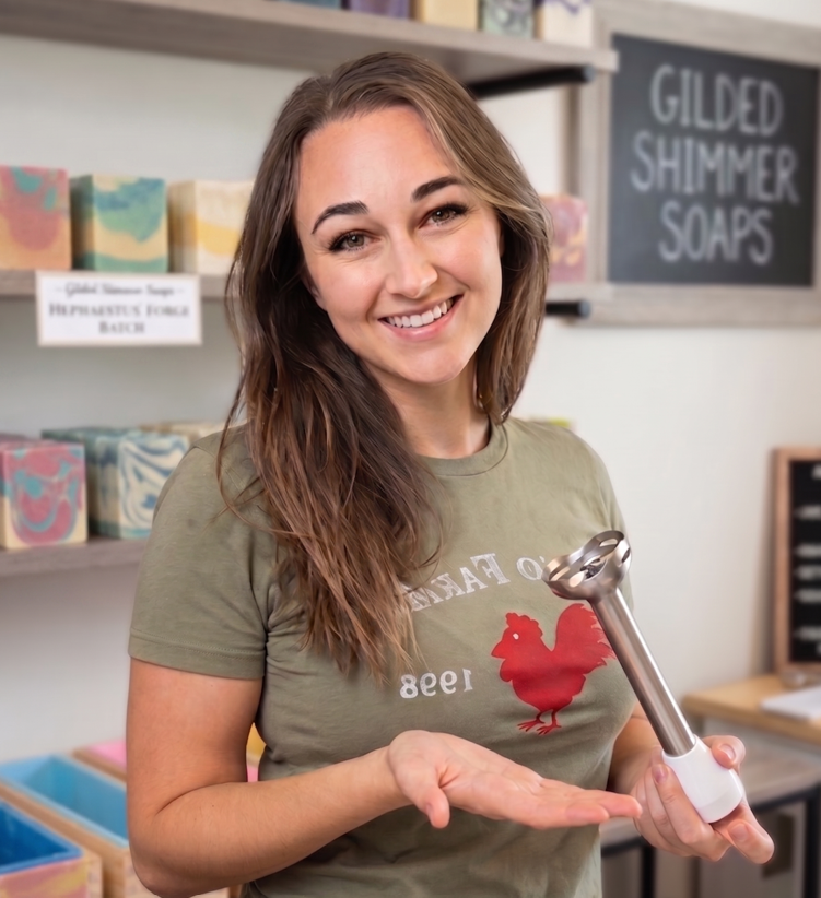 Woman holding a soap-making tool in a soap-making studio with shelves of soap products in the background.